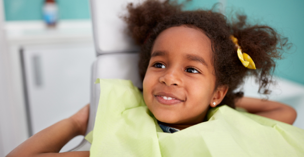 Young child smiling in bright yellow shirt at pediatric dental office with turquoise background.
