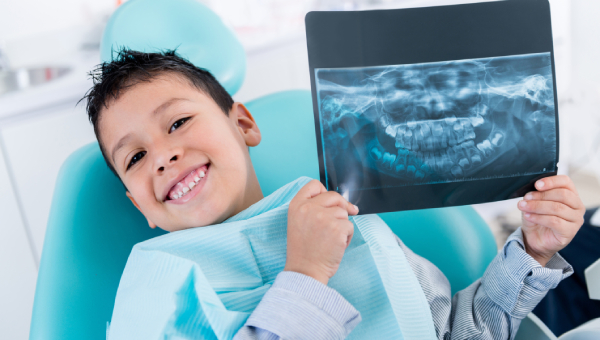 Young child in dental chair wearing bib holding X-ray image, smiling at camera.