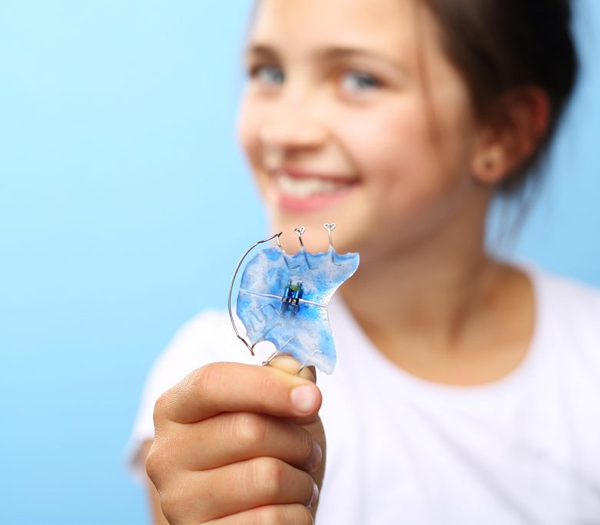 Child holding clear orthodontic retainer against blue background, smiling.