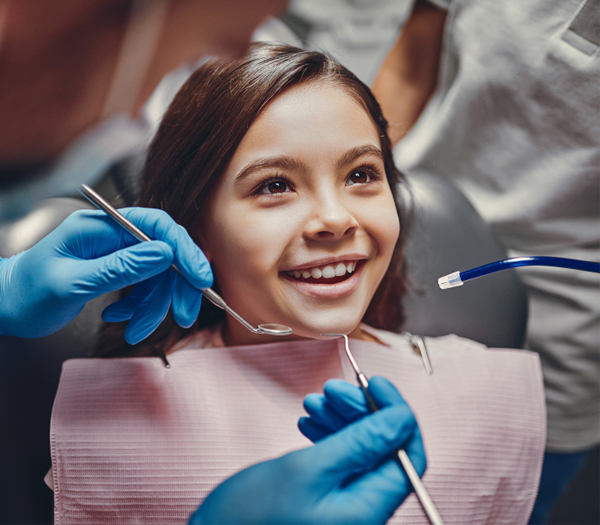 Young girl smiling in dental chair during pediatric dental examination with dentist holding instruments.