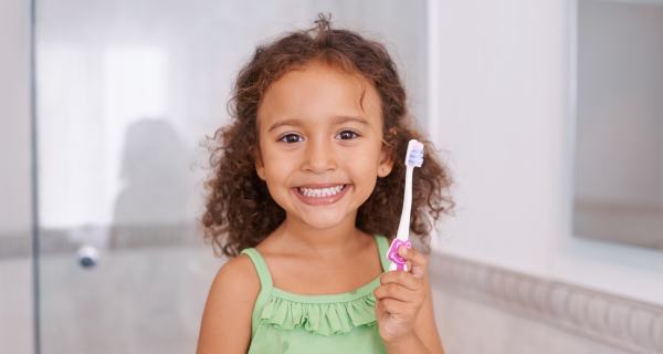 Smiling young girl with curly hair holding toothbrush in bright bathroom setting.