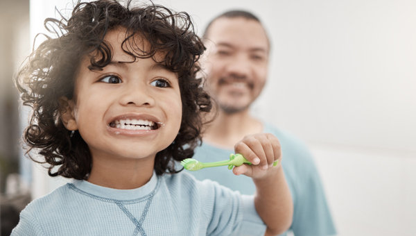 Young child brushing teeth with father supervising in bright bathroom setting.