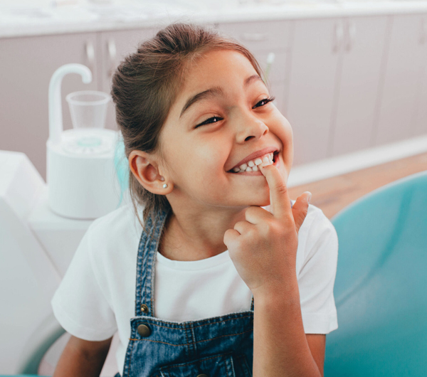 Young girl smiling in dental office, touching her tooth, wearing blue apron.