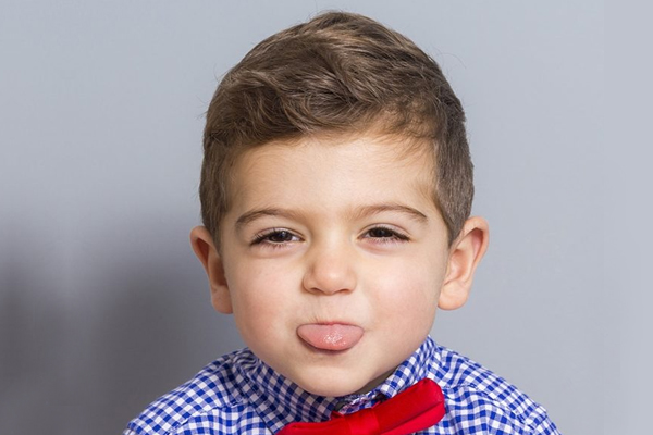 Young boy in blue checkered shirt and red bow tie smiling at camera against gray background.